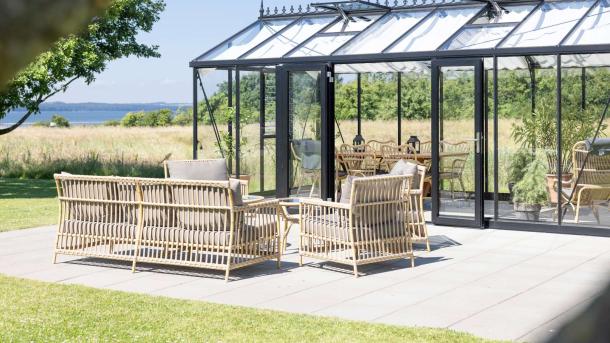 Væksthus eller orangeri, der er indrettet med kurvemøbler, ses udefra med hav i baggrunden